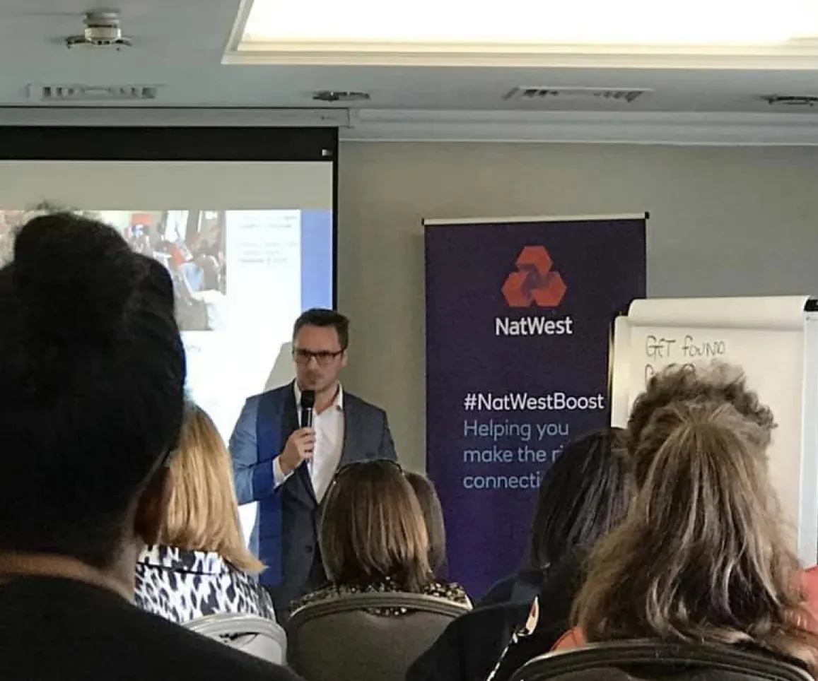 Dr Lewis Haydon speaking with a microphone to an audience of business owners at NatWest Boost